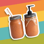 A rust colour mason jar toothbrush holder and  soap dispenser combo on a colorful striped background.