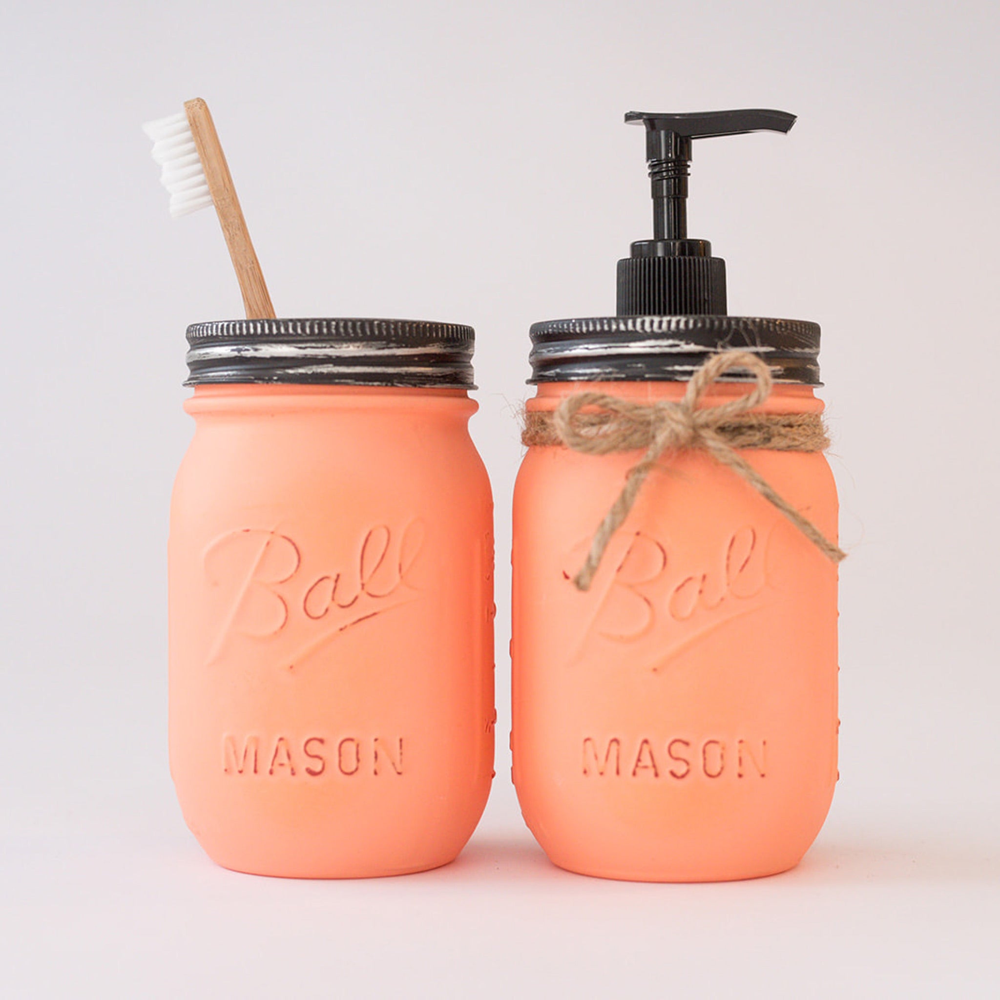 Two coral-colored mason jars with a toothbrush and pump dispenser on a light gray background.