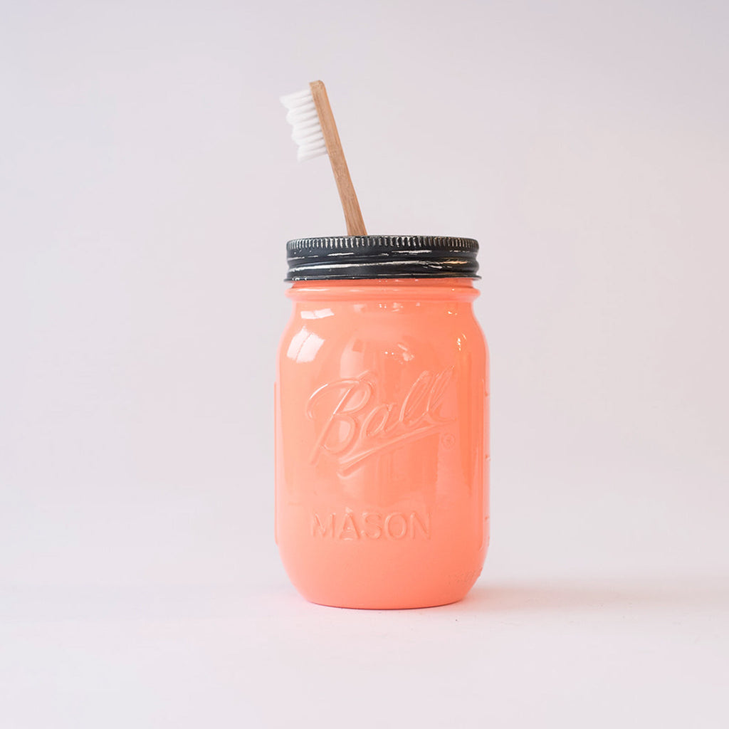 A coral mason jar with metal lid and toothbrush in it on a white background.