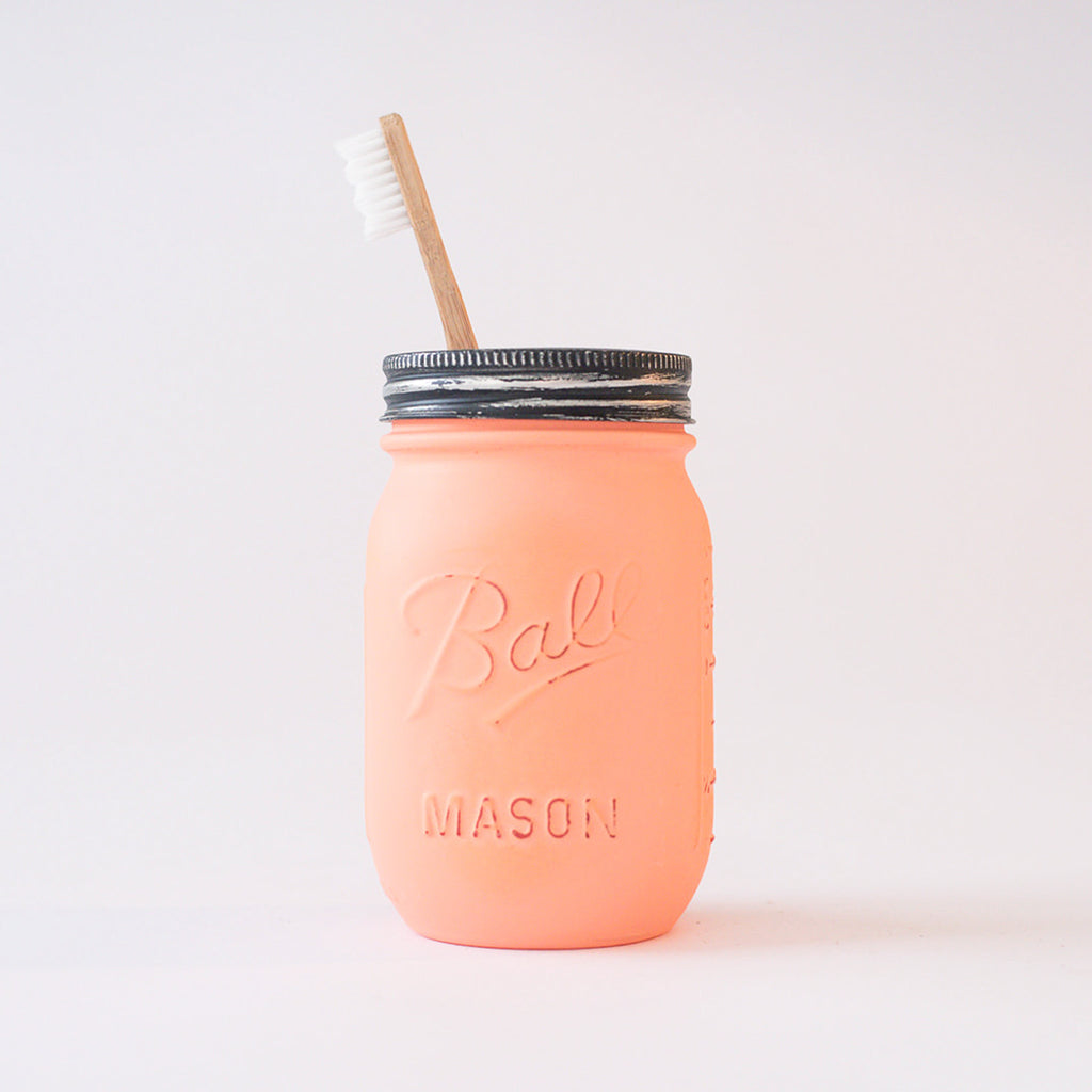 A coral-colored mason jar with a toothbrush in it on a white background.